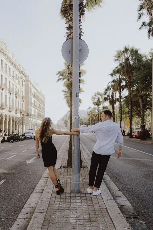 Couple playfully holding hands around a lamppost on a palm-lined city street during an engagement photoshoot.