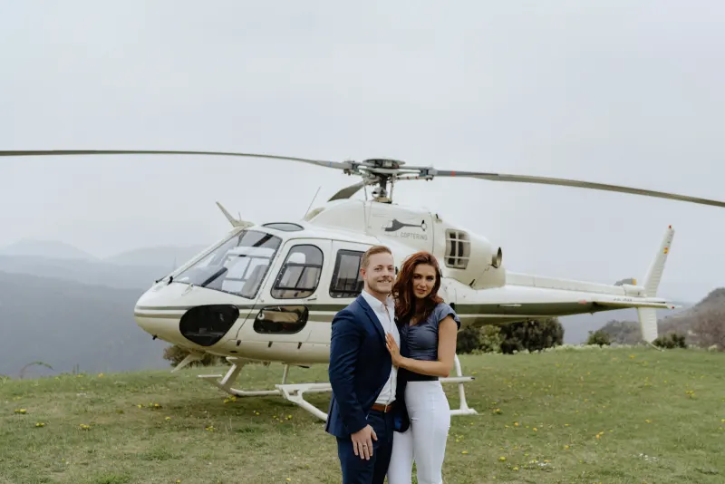Couple poses in front of a helicopter on a scenic hillside, capturing excitement before their surprise proposal photoshoot.
