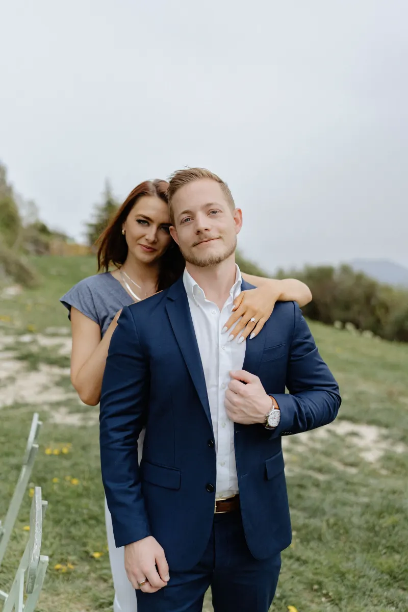 Couple poses together on a grassy hillside, showing off her engagement ring after a surprise proposal photoshoot.
