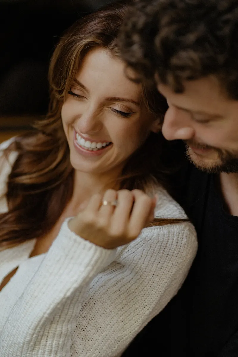 Couple shares a candid laugh as she admires her engagement ring during a cozy couples & engagements photoshoot.