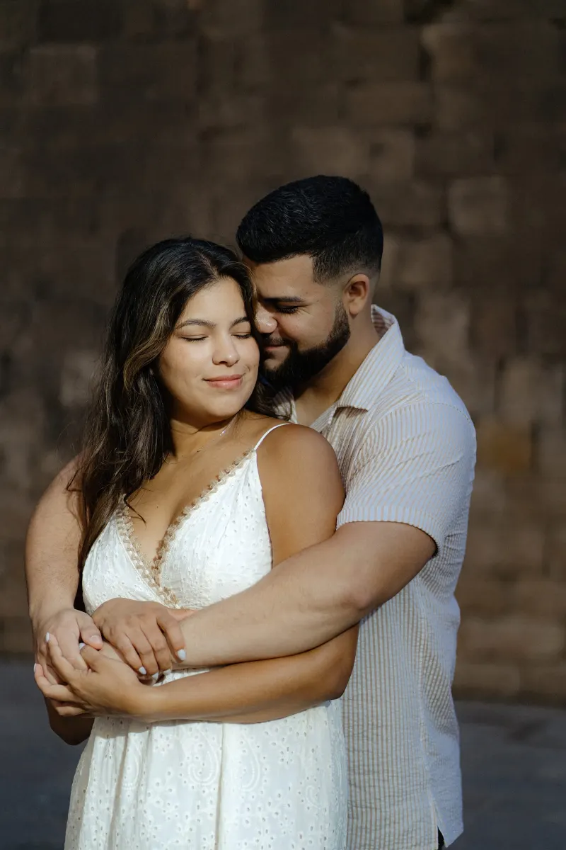 Couple shares a gentle embrace against a rustic stone wall during their couples & engagements photoshoot.