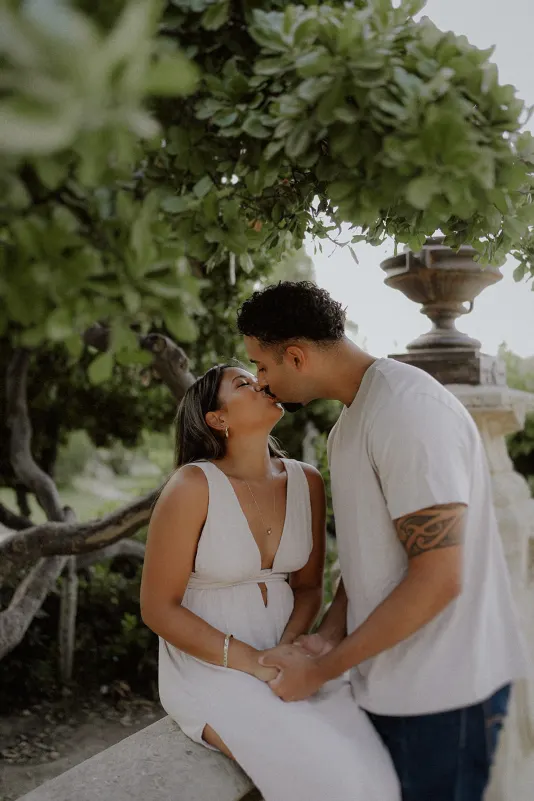 Couple shares a gentle kiss beneath lush greenery during an intimate engagement photoshoot in a garden setting.