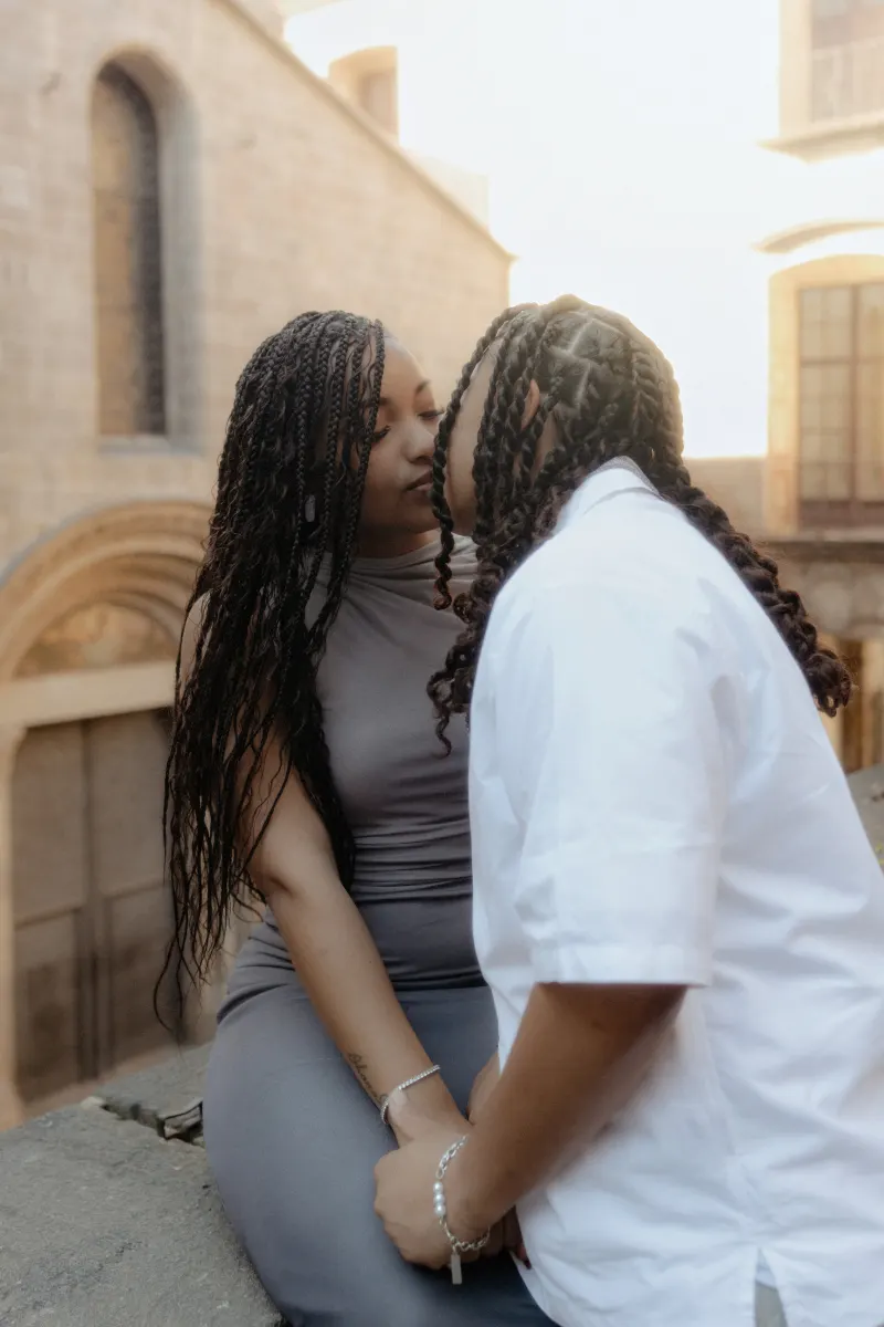 Couple shares a gentle kiss, holding hands on a stone ledge during a surprise proposal photoshoot in Barcelona.