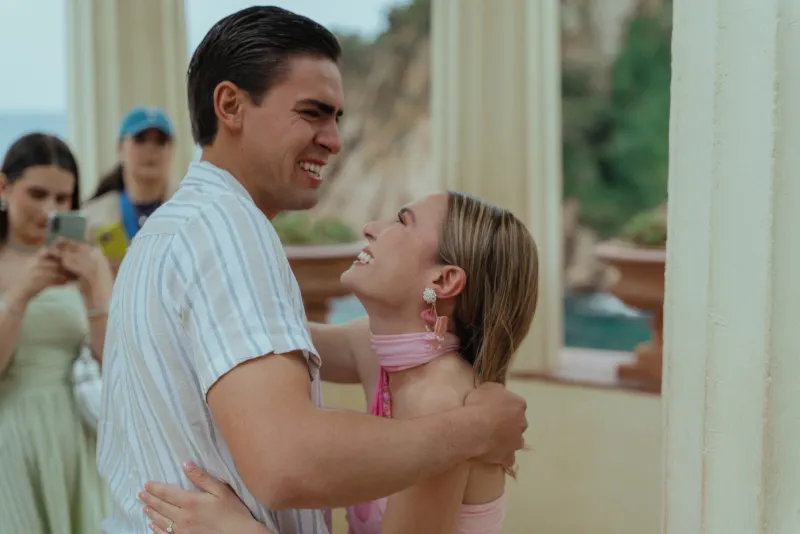 Couple shares a joyful embrace after a seaside proposal, captured during a surprise proposal photoshoot with friends nearby.