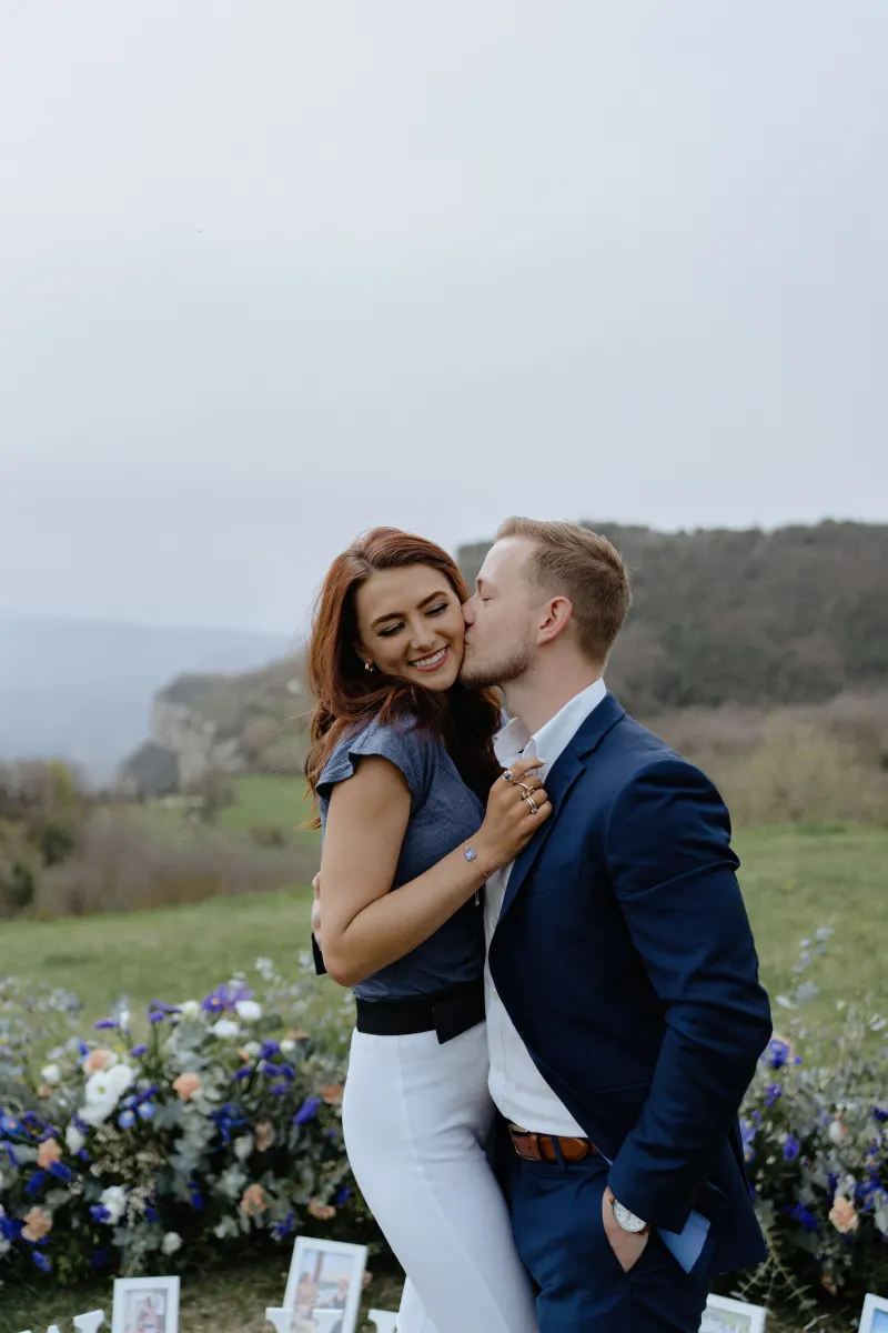 Couple shares a joyful embrace and kiss on a hillside, celebrating their engagement during a surprise proposal photoshoot.