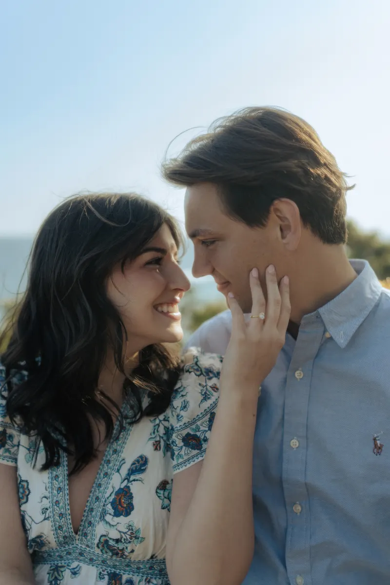 Couple shares a joyful embrace by the sea, her engagement ring visible during a surprise proposal photoshoot.