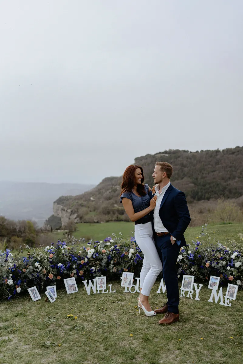 Couple shares a joyful embrace on a flower-lined hillside, celebrating their engagement during a surprise proposal photoshoot