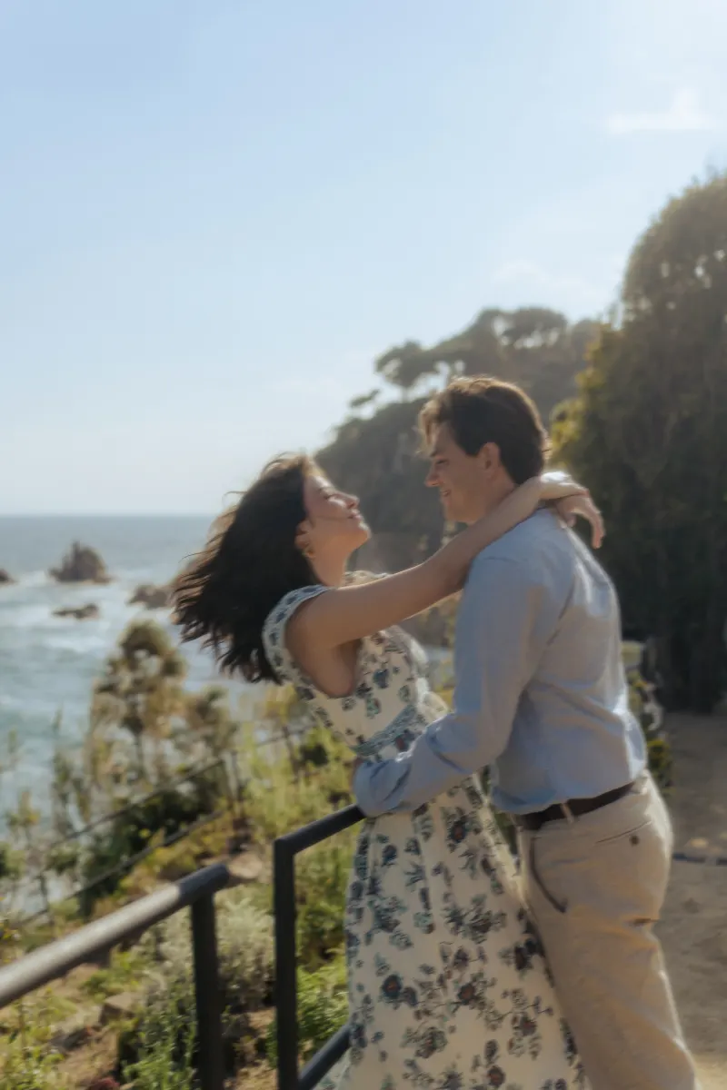 Couple shares a joyful embrace on a sunlit coastal path, captured during a surprise proposal photoshoot.
