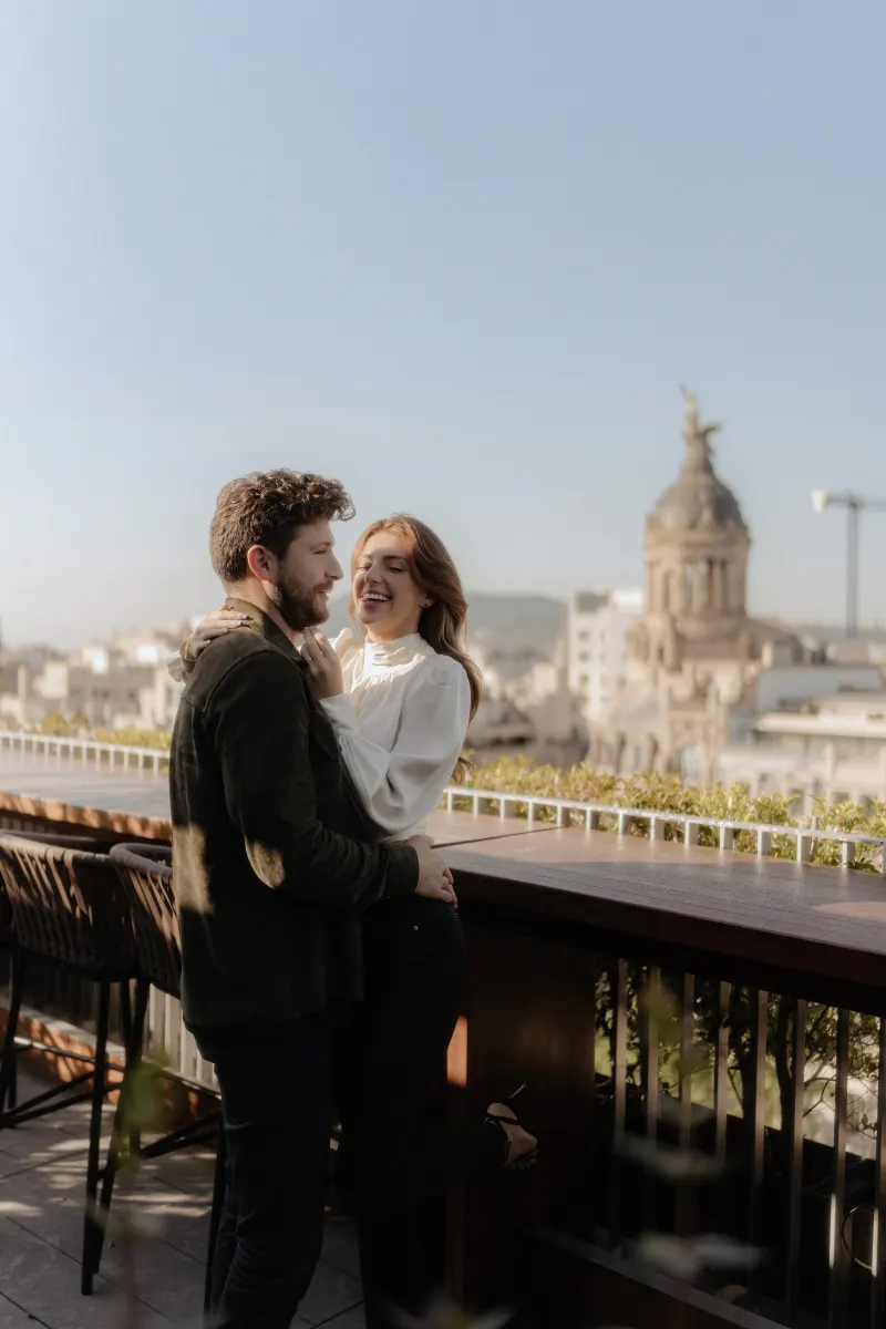 Couple shares a joyful embrace on a sunlit rooftop during a couples & engagements photoshoot with city landmarks in view.