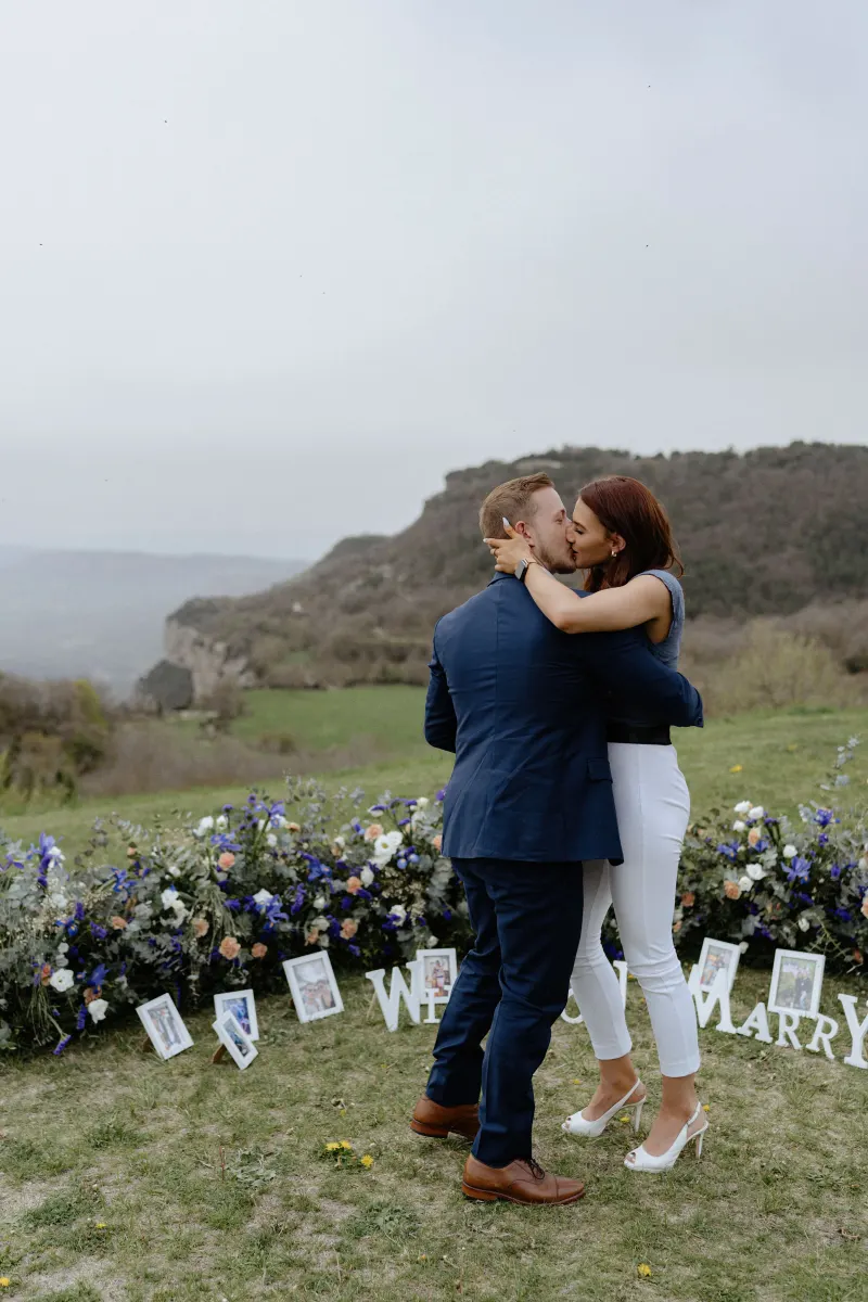 Couple shares a joyful kiss on a flower-lined hillside after a surprise proposal, captured during a professional photoshoot.