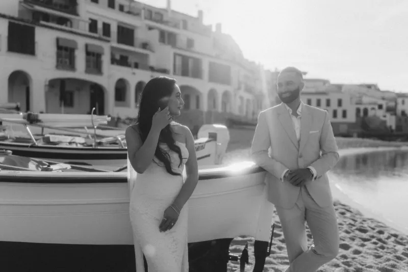 Couple shares a joyful moment by boats on a sunlit beach during a surprise proposal photoshoot in a coastal village.