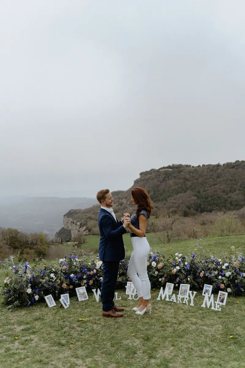 Couple shares a joyful moment on a flower-lined hillside, celebrating their engagement during a surprise proposal photoshoot.