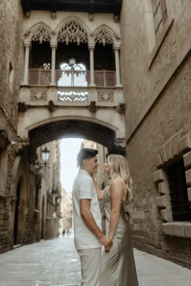 Couple shares a loving gaze while holding hands beneath Barcelona’s Gothic bridge during a Vacation Portraits photoshoot.