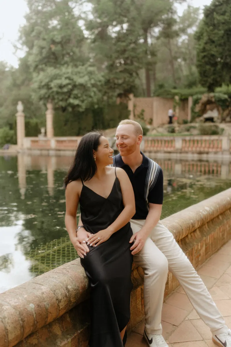 Couple shares a loving smile while seated by a tranquil pond during a surprise proposal photoshoot in a garden.