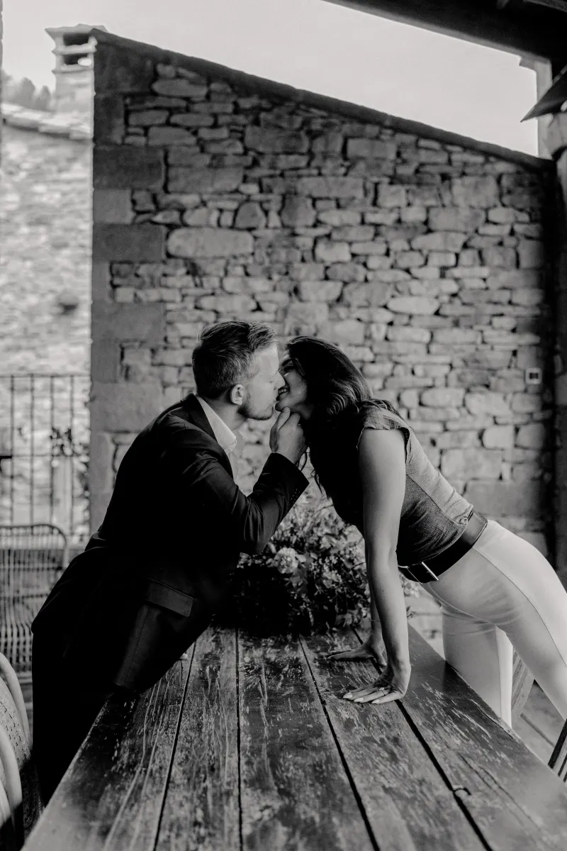 Couple shares a playful kiss across a rustic wooden table, celebrating their engagement after a surprise proposal session.