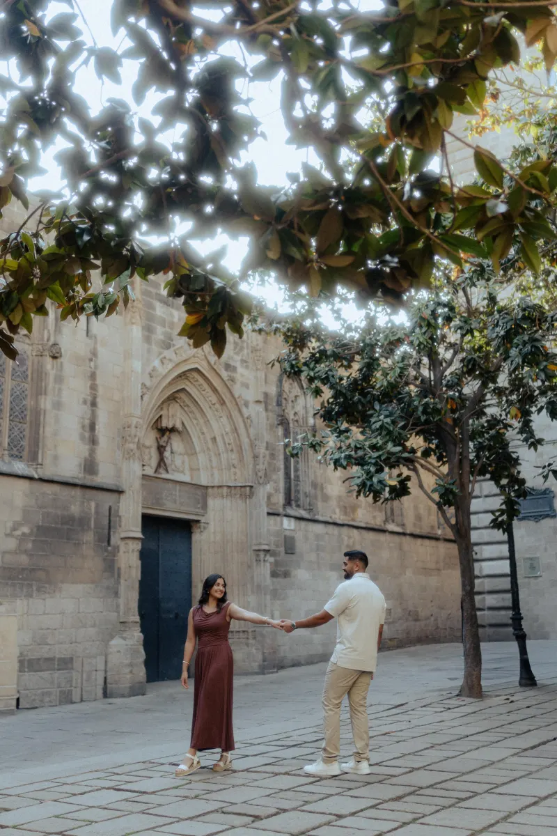 Couple shares a playful moment, holding hands beneath leafy trees during a couples & engagements photoshoot in a historic squ