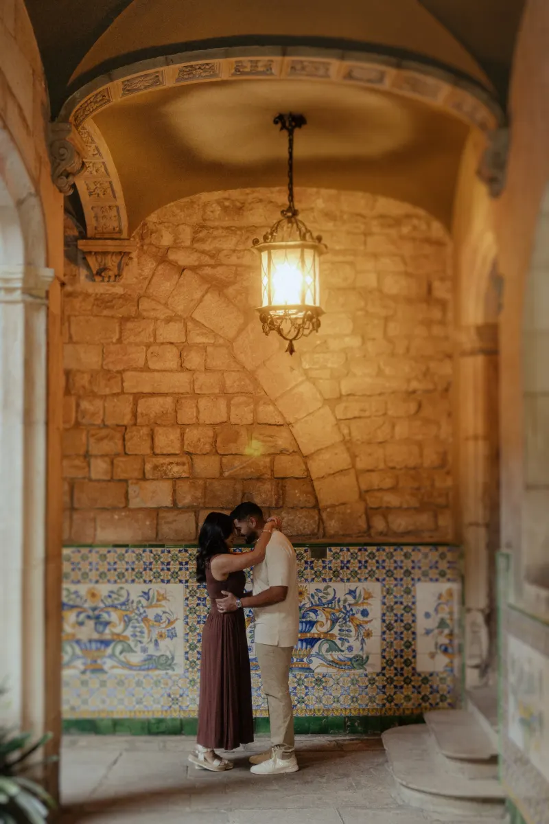 Couple shares a quiet embrace beneath a glowing lantern during their engagement photoshoot in a historic tiled corridor.