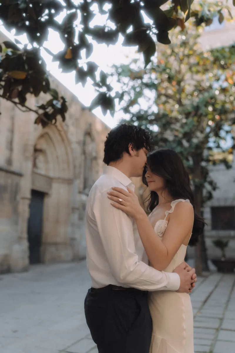Couple shares a quiet embrace under leafy trees in a historic courtyard during a couples & engagements photoshoot.