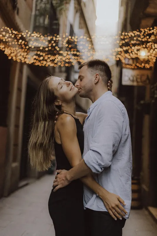 Couple shares a romantic embrace under string lights in a narrow city street during a couples & engagements photoshoot.