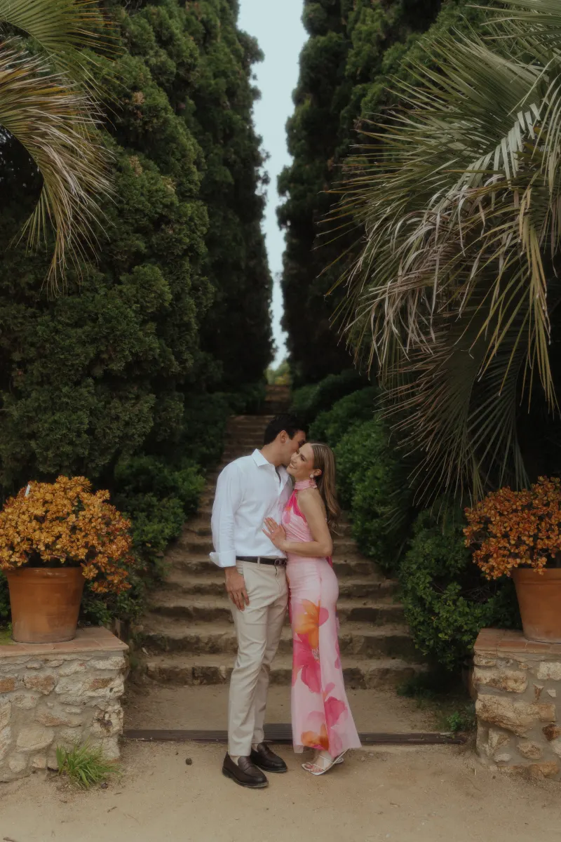 Couple shares a romantic kiss on a garden path, captured during a surprise proposal photoshoot among lush greenery.