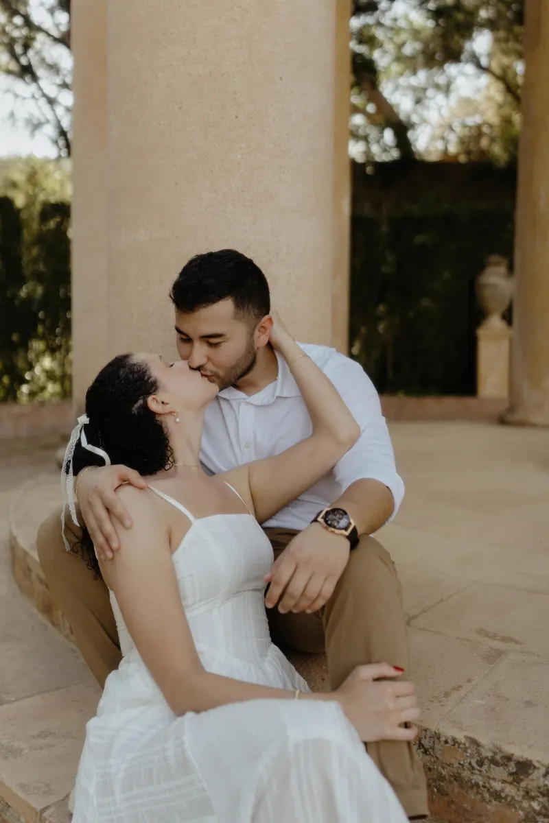 Couple shares a romantic kiss while seated on stone steps during a couples & engagements photoshoot at a garden pavilion.