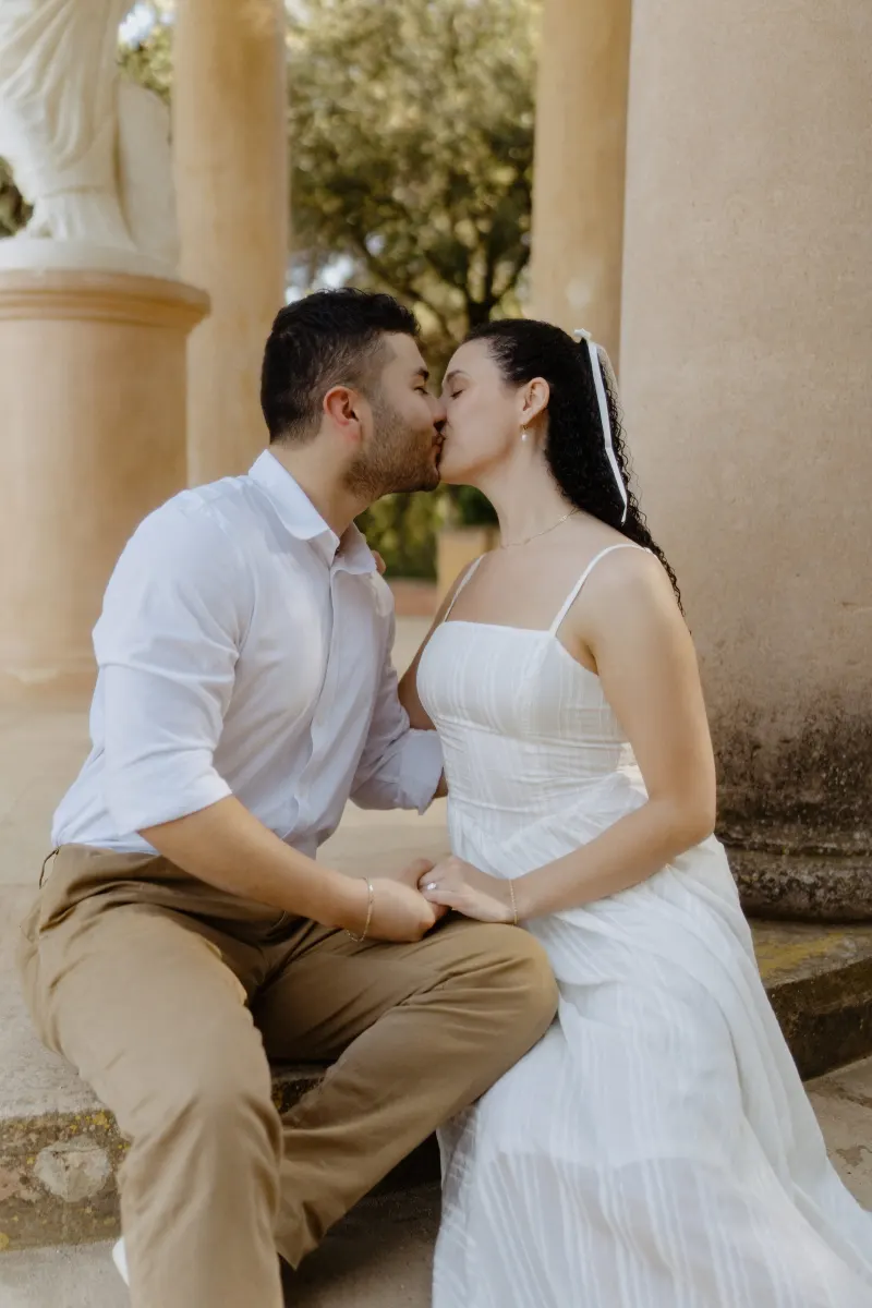 Couple shares a sweet kiss while seated on stone steps between columns during a couples & engagements photoshoot.