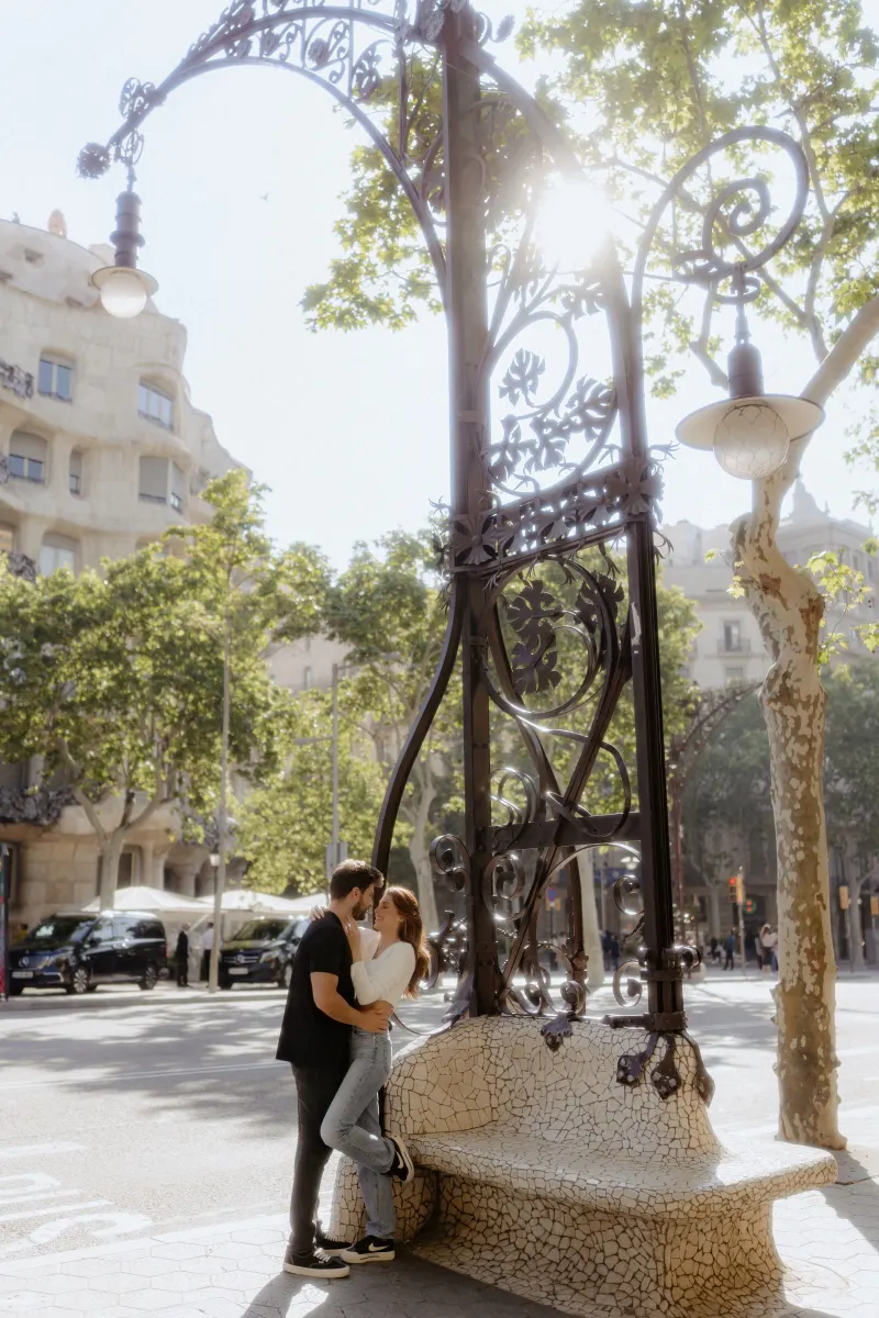 Couple shares a tender embrace under ornate ironwork on a sunlit city street during a couples & engagements photoshoot.