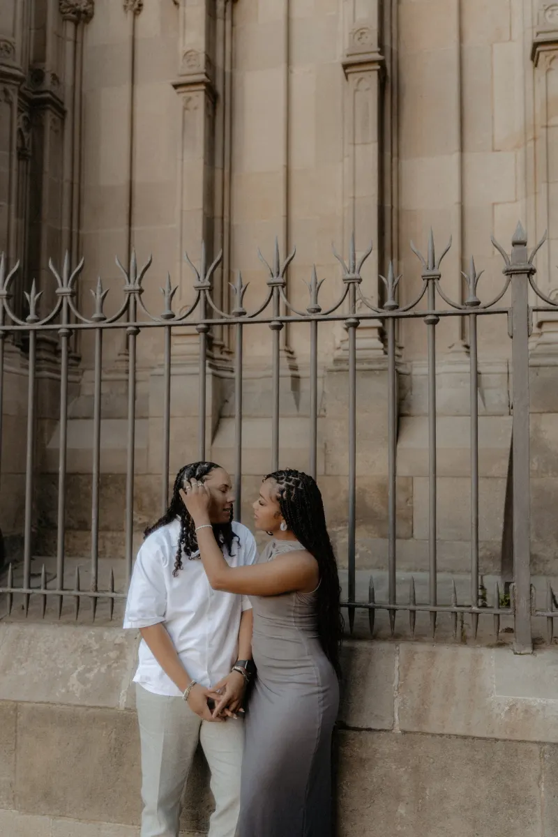 Couple shares a tender gaze and holds hands by the cathedral fence during a surprise proposal photoshoot in Barcelona.