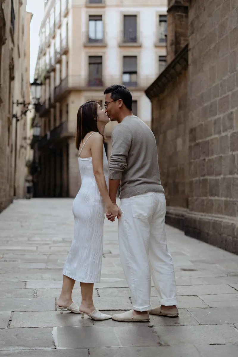 Couple shares a tender kiss while holding hands during a Vacation Portraits photoshoot in a quiet Barcelona street.