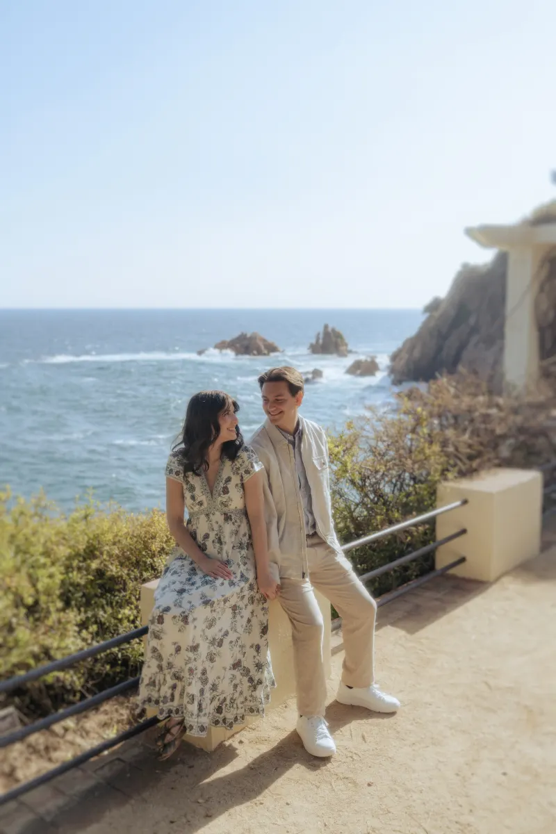 Couple shares a tender moment by the ocean, holding hands during a surprise proposal photoshoot on a sunny day.