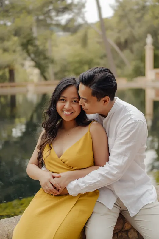 Couple shares a warm embrace by a tranquil pond, smiling during their couples & engagements photoshoot in the park.