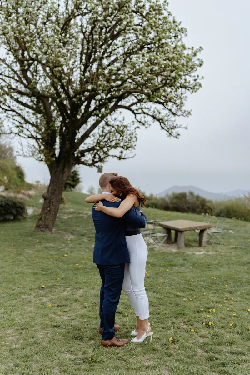 Couple shares an emotional embrace under a blooming tree after a countryside surprise proposal photoshoot.