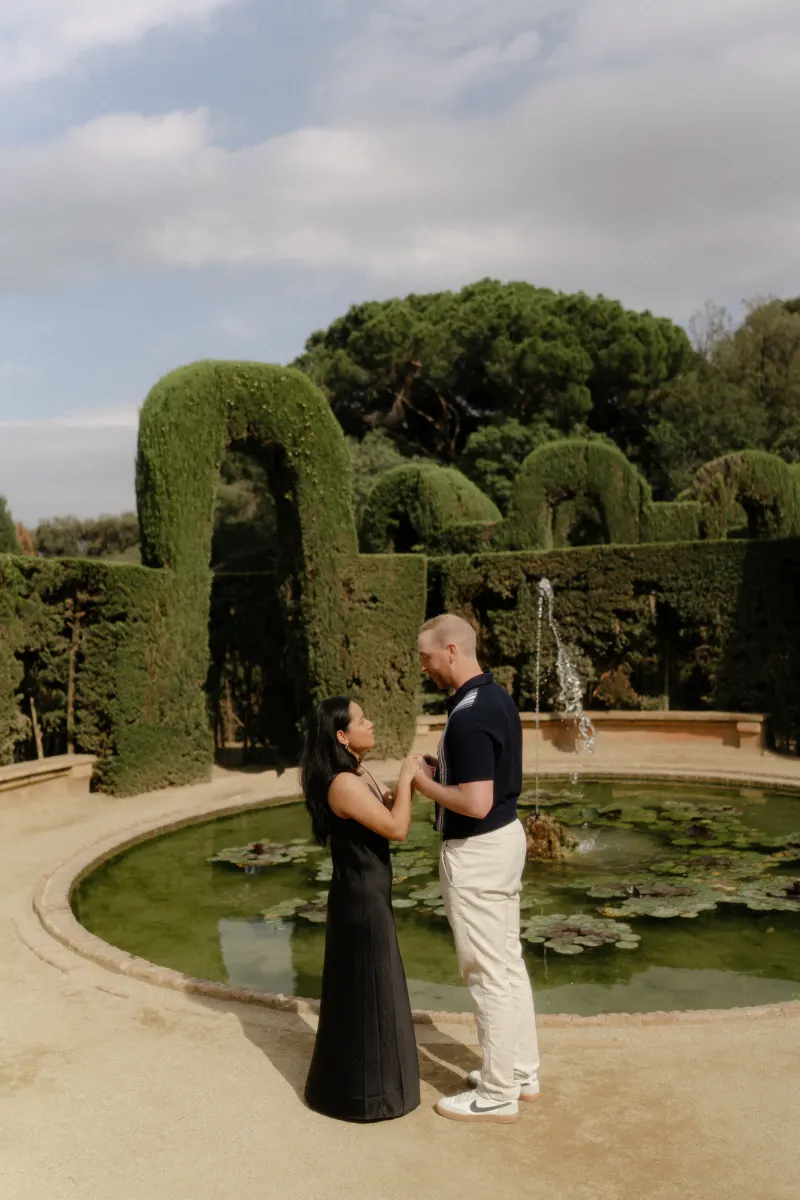 Couple shares an emotional moment by a garden fountain during a surprise proposal photoshoot in a lush park setting.
