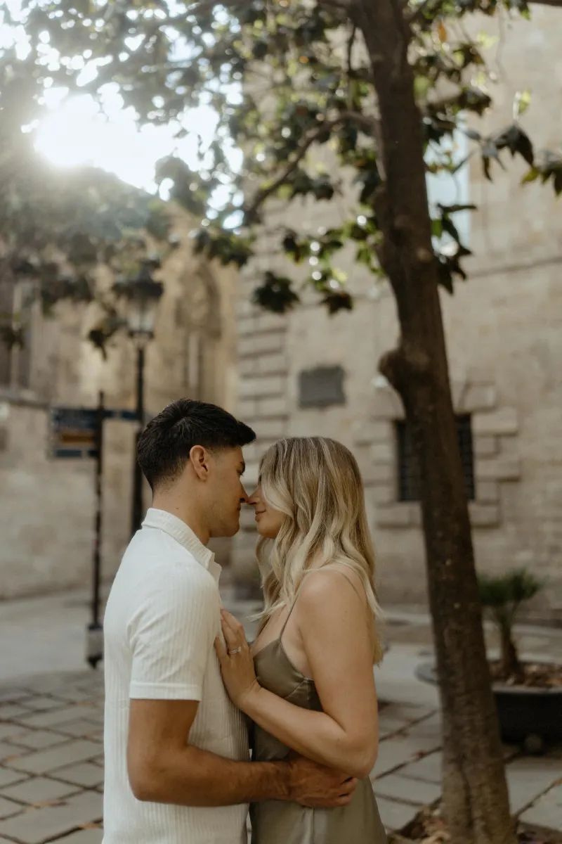Couple sharing a gentle forehead touch beneath sunlit trees in Barcelona during a Vacation Portraits photoshoot.