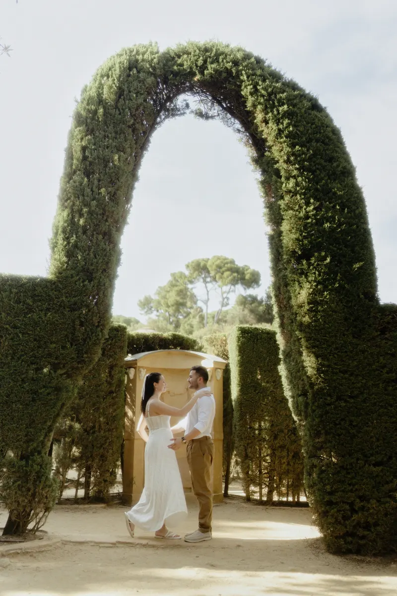 Couple sharing a joyful moment beneath a tall garden arch during an engagement photoshoot in a sunlit park.