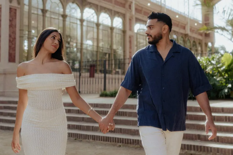 Couple sharing a loving gaze while walking hand in hand outside a sunlit conservatory during an engagement photoshoot.