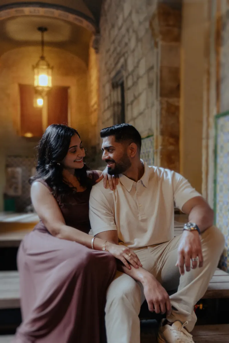Couple sharing a loving smile while seated together during a couples & engagements photoshoot in a warmly lit historic hallwa