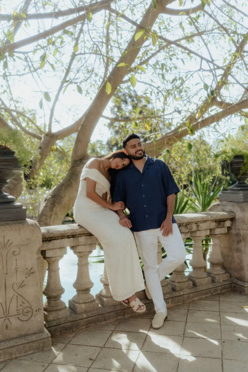 Couple sharing a peaceful moment by a stone railing under sun-dappled trees during an outdoor engagement photoshoot.