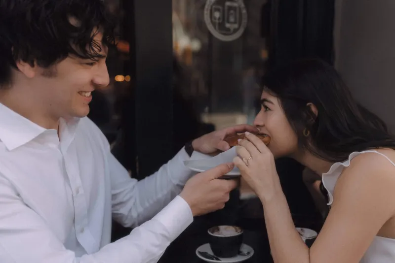 Couple sharing a playful bite of pastry at a café, laughing together during their couples & engagements photoshoot.