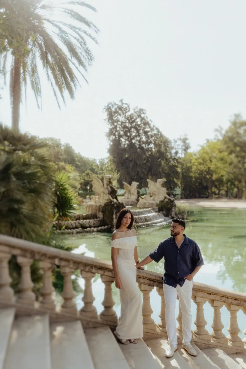 Couple sharing a quiet, romantic moment by a sunlit fountain during an engagement photoshoot in a lush garden setting.