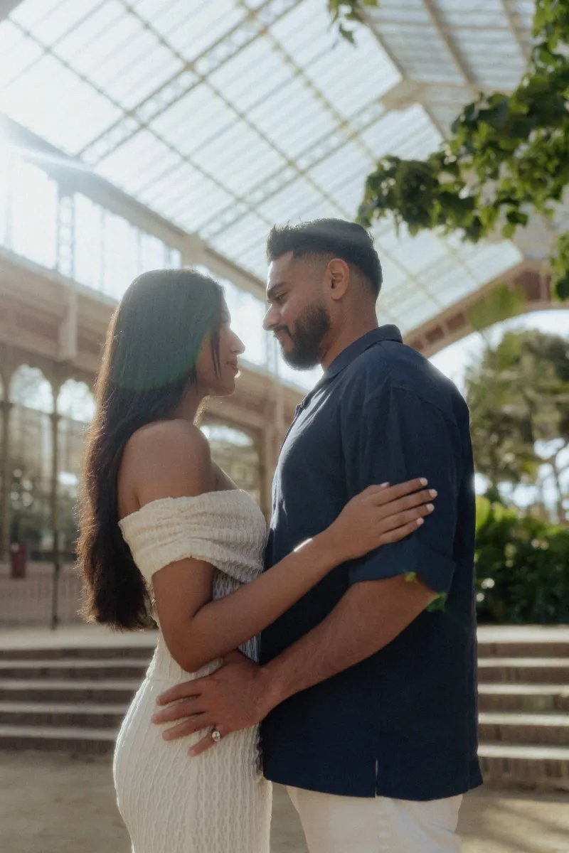 Couple sharing an intimate embrace and smiling softly during a couples photoshoot in a sunlit glass conservatory.