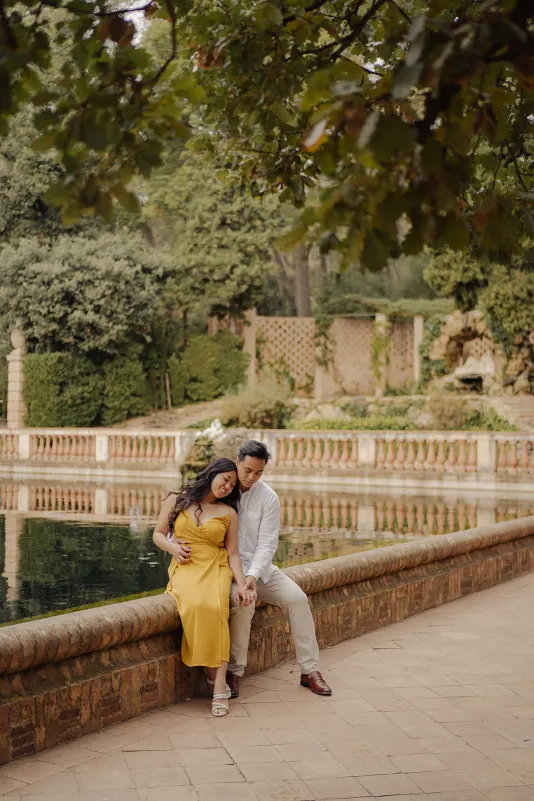 Couple sits close by a tranquil pond, sharing a peaceful moment during a couples & engagements photoshoot in the park.