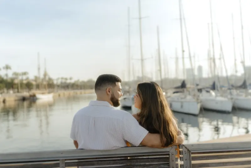 Couple sits close on a marina bench, sharing a loving gaze during a couples & engagements photoshoot by the water.