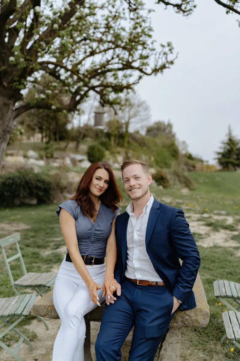 Couple sits close on a stone bench, holding hands and smiling during a countryside surprise proposal photoshoot.