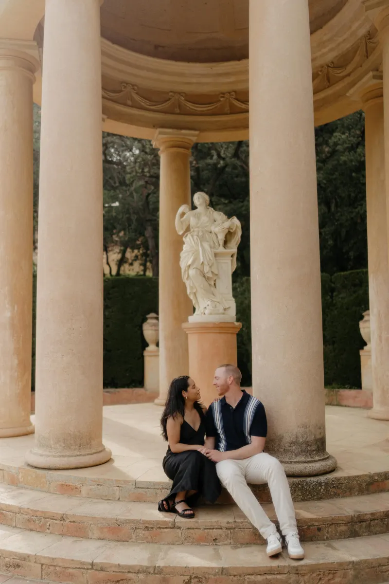 Couple sits close on stone steps of a classical pavilion, sharing a tender moment during a surprise proposal photoshoot.