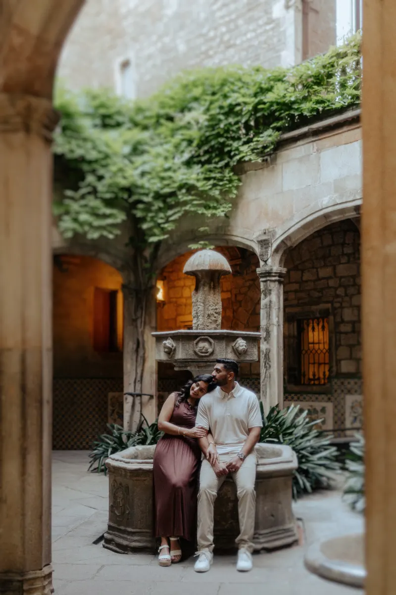 Couple sits close together on a stone fountain, sharing a quiet moment during a couples & engagements photoshoot in a courtya