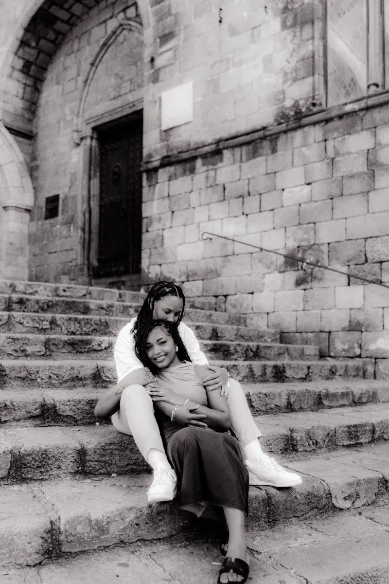 Couple sits embraced on historic stone steps in Barcelona, sharing a joyful moment during their surprise proposal photoshoot.