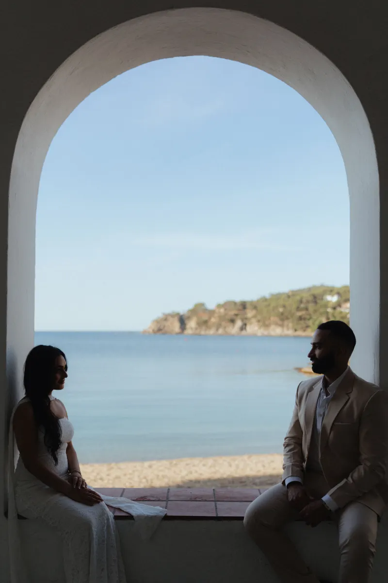 Couple sits facing each other by the sea after a surprise proposal photoshoot, framed by a sunlit archway.