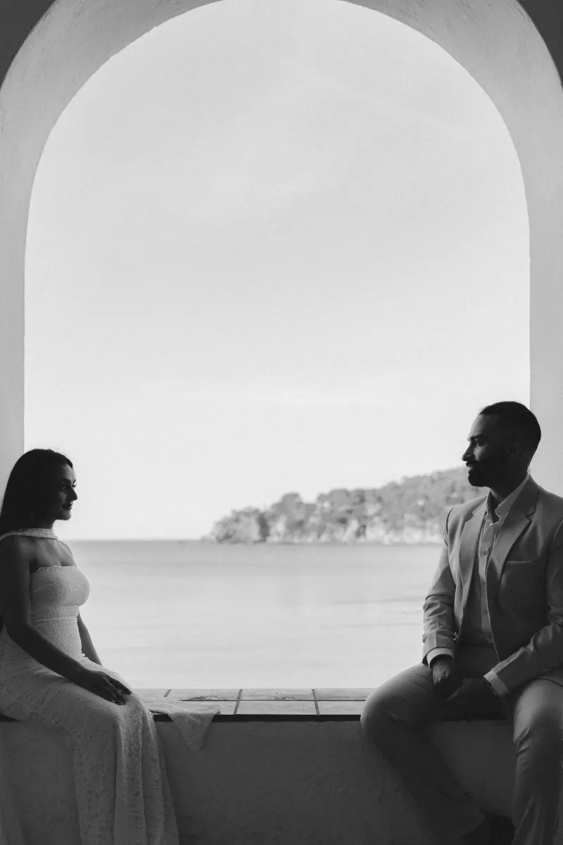 Couple sits facing each other in a romantic surprise proposal photoshoot by the sea, framed by an arched window.