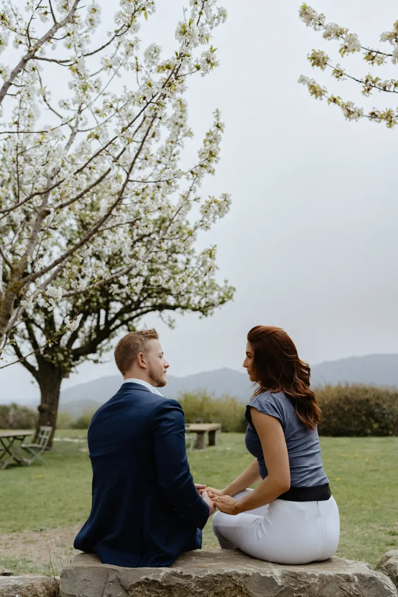 Couple sits hand in hand beneath blooming trees, sharing a quiet moment before their countryside surprise proposal session.